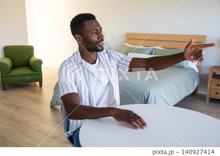 African american man sitting in bedroom at white table pointing toward bed frame with bedding African american man sitting in bedroom at white table pointing toward bed frame with bedding 140927414