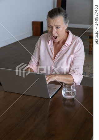 Woman typing on laptop at home on table with water glass looking surprised Woman typing on laptop at home on table with water glass looking surprised 140927453