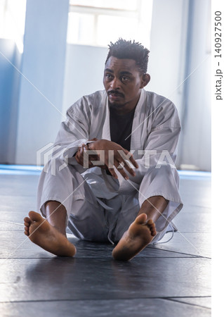 African american man stretching wrist, sitting on padded mat in dojo wearing white gi African american man stretching wrist, sitting on padded mat in dojo wearing white gi 140927500