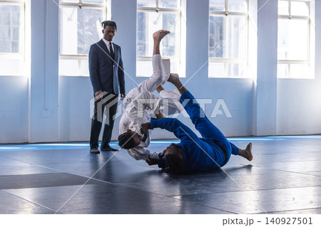 African american male fighters in gis executing grappling move on mats while referee observing 140927501
