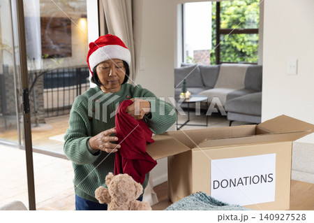 Senior Asian woman wearing Santa hat placing red garment into donation box at living room table 140927528