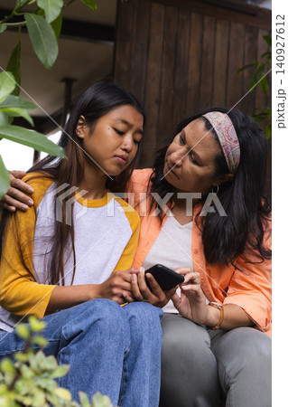 Mother and daughter sitting on wooden porch bench sharing smartphone and leaning into leafy plants Mother and daughter sitting on wooden porch bench sharing smartphone and leaning into leafy plants 140927612