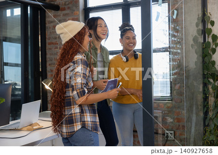 Diverse female coworkers brainstorming at glass partition in meeting area with tablet and marker Diverse female coworkers brainstorming at glass partition in meeting area with tablet and marker 140927658