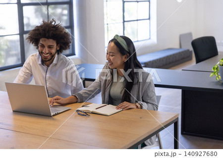 Diverse coworkers working at office desk with laptop, notebook, pen, eyeglasses, potted plant Diverse coworkers working at office desk with laptop, notebook, pen, eyeglasses, potted plant 140927680