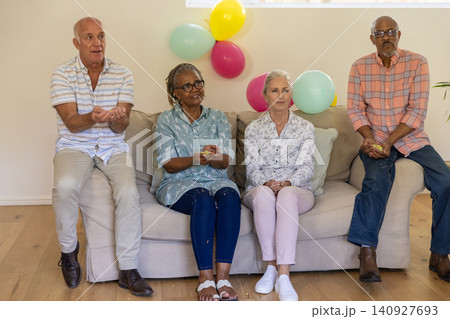 Senior friends sitting on beige couch in living room holding confetti near plant, pastel balloons Senior friends sitting on beige couch in living room holding confetti near plant, pastel balloons 140927693