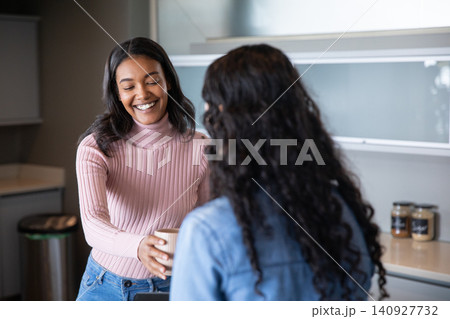 Diverse female coworkers exchanging coffee mug in office kitchenette with jars, trash bin Diverse female coworkers exchanging coffee mug in office kitchenette with jars, trash bin 140927732