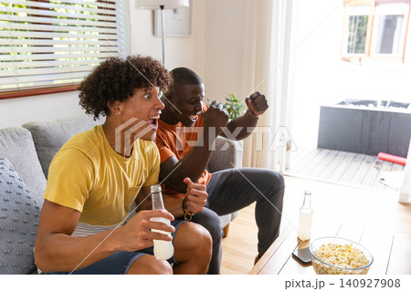 Diverse male friends cheering on sofa at home holding beverage bottles near popcorn Diverse male friends cheering on sofa at home holding beverage bottles near popcorn 140927908