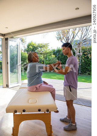 Diverse therapist and patient performing seated arm stretch on therapy table by glass doors Diverse therapist and patient performing seated arm stretch on therapy table by glass doors 140927956
