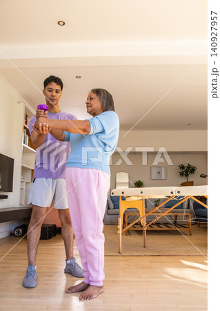 Senior woman and male trainer raising purple dumbbell beside therapy table in home area Senior woman and male trainer raising purple dumbbell beside therapy table in home area 140927957