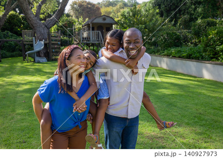 Diverse family carrying daughters piggyback while walking across backyard lawn Diverse family carrying daughters piggyback while walking across backyard lawn 140927973