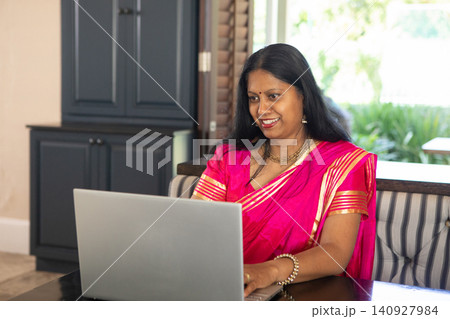 Mature indian woman wearing pink saree with gold jewelry and working on laptop at home office 140927984