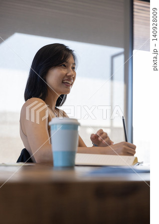 Asian woman writing in notebook at table in study room with coffee cup blue sleeve Asian woman writing in notebook at table in study room with coffee cup blue sleeve 140928009