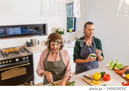 Senior female friends chopping cucumber, mixing greens at kitchen island with cutting boards Senior female friends chopping cucumber, mixing greens at kitchen island with cutting boards 140928038