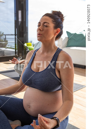Pregnant woman meditating with partner on yoga mats in living room with plants, wearing smartwatch Pregnant woman meditating with partner on yoga mats in living room with plants, wearing smartwatch 140928073