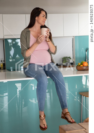 Mid adult pregnant woman sitting on kitchen island holding mug beside fruit bowl while gazing left Mid adult pregnant woman sitting on kitchen island holding mug beside fruit bowl while gazing left 140928075