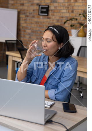 Asian woman sipping water from glass, typing on laptop and checking smartphone at office desk Asian woman sipping water from glass, typing on laptop and checking smartphone at office desk 140928172