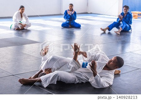 Diverse martial arts students grappling on padded mats in dojo wearing gis and belts Diverse martial arts students grappling on padded mats in dojo wearing gis and belts 140928213
