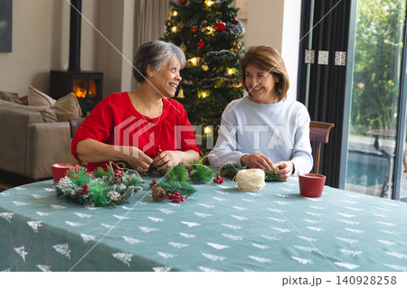 Senior, middle-aged women friends crafting festive wreaths in living room with pine cones and twine Senior, middle-aged women friends crafting festive wreaths in living room with pine cones and twine 140928258