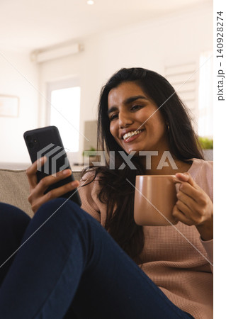 Indian woman smiling and looking at smartphone while holding ceramic mug on sofa in living room Indian woman smiling and looking at smartphone while holding ceramic mug on sofa in living room 140928274