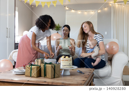 Diverse female friends with infant gathering around sofa at home displaying cake, gifts Diverse female friends with infant gathering around sofa at home displaying cake, gifts 140928280