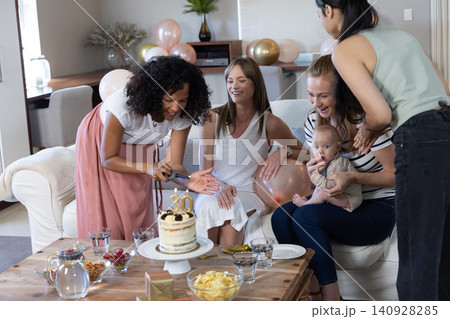 Diverse female friends cutting birthday cake on wooden coffee table in living room, balloons Diverse female friends cutting birthday cake on wooden coffee table in living room, balloons 140928285