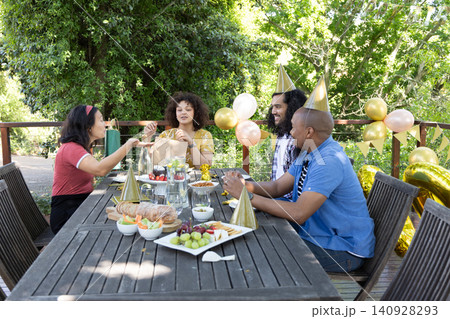 Diverse friends sitting around table on deck sharing gift bag, sampling pastries amid balloons Diverse friends sitting around table on deck sharing gift bag, sampling pastries amid balloons 140928293
