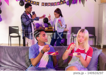 Diverse friends celebrating birthday on grey couch with banner balloons holding cake, pink cups 140928297
