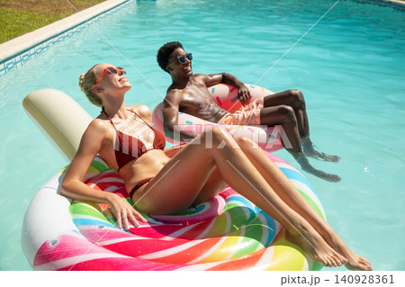Diverse couple lounging in swimwear on spiral, donut floats at backyard pool wearing sunglasses 140928361