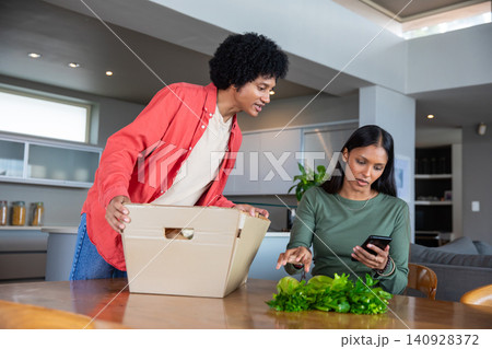 Diverse couple unpacking cardboard box and examining leafy greens, using smartphone in kitchen Diverse couple unpacking cardboard box and examining leafy greens, using smartphone in kitchen 140928372