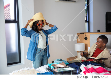 African American couple packing suitcase with straw hat, red sunglasses in bedroom holding notebook African American couple packing suitcase with straw hat, red sunglasses in bedroom holding notebook 140928398