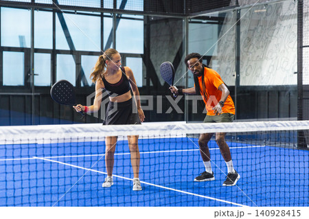Diverse sports partners standing behind tennis net on padel court in sports hall holding rackets Diverse sports partners standing behind tennis net on padel court in sports hall holding rackets 140928415
