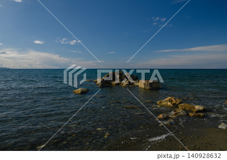 Serene coastline with gentle waves and rugged rocks under a clear blue sky at midday Serene coastline with gentle waves and rugged rocks under a clear blue sky at midday 140928632