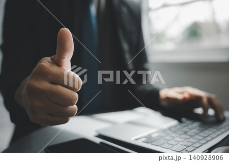 A closeup of a man hand pose with a success sign gesture, the businessman gives a thumbs up sign for approval while working on a laptop at his modern office desk 140928906