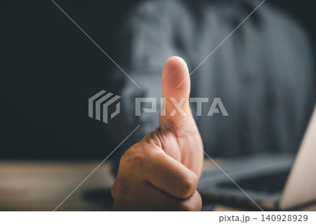 Close-up of a hand giving a thumbs-up, blurry background with a laptop out of focus. Close-up of a hand giving a thumbs-up, blurry background with a laptop out of focus. 140928929