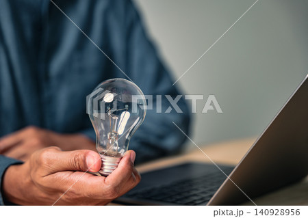 A man makes a creative hand gesture to pose with a lightbulb, a sign of a new idea while he is posing for business inspiration at his desk with a computer 140928956