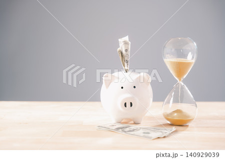 White piggy bank holds money, symbolizing saving money concept, time represented by elegant hourglass glass with sand flowing for business investment, future retirement planning White piggy bank holds money, symbolizing saving money concept, time represented by elegant hourglass glass with sand flowing for business investment, future retirement planning 140929039