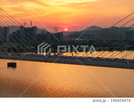 Aerial view captures a golden hour light bathes a modern city skyline and cable-stayed bridge spanning a calm river at dusk Aerial view captures a golden hour light bathes a modern city skyline and cable-stayed bridge spanning a calm river at dusk 140929478