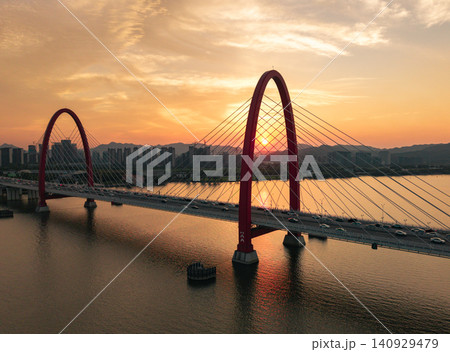 Aerial view captures golden sunset framing a prominent red arch bridge spanning a wide river with city skyline and mountains at dusk Aerial view captures golden sunset framing a prominent red arch bridge spanning a wide river with city skyline and mountains at dusk 140929479