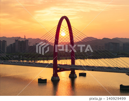 Aerial view captures golden sunset framing a prominent red arch bridge spanning a wide river with city skyline and mountains at dusk Aerial view captures golden sunset framing a prominent red arch bridge spanning a wide river with city skyline and mountains at dusk 140929499