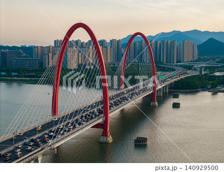 Aerial view captures red arched cable bridge filled with vehicles crosses a wide river flanked by urban buildings and distant mountains Aerial view captures red arched cable bridge filled with vehicles crosses a wide river flanked by urban buildings and distant mountains 140929500