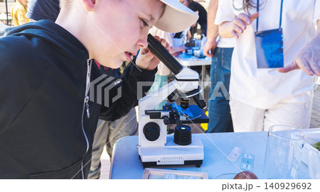 Curious boy looking through a microscope at an outdoor science fair. Enthusiastic kid exploring biology sample during an educational laboratory festival. 140929692