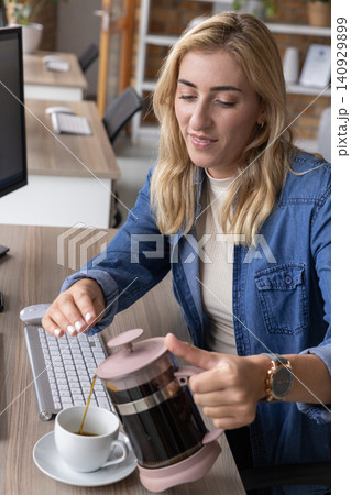 Mid-adult woman in denim shirt pouring coffee from pink French press into cup at office desk Mid-adult woman in denim shirt pouring coffee from pink French press into cup at office desk 140929899