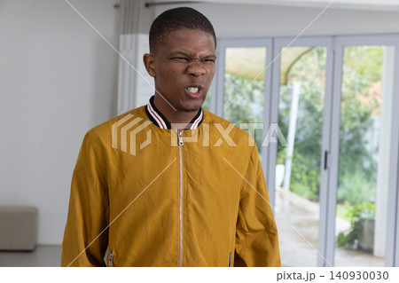 African American man grimacing in living room by sliding glass doors wearing mustard jacket 140930030