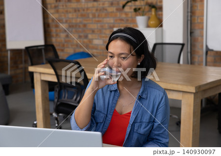 Asian woman wearing denim shirt sitting at wooden meeting table holding glass and checking laptop 140930097