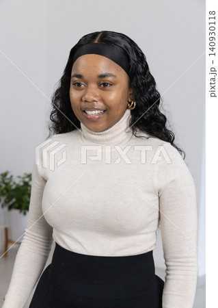 African American woman posing smiling in studio wearing cream-sweater and gold-hoop near plant 140930118
