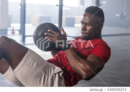 African american man gripping black medicine ball, leaning back on gym mat near free-weight rack 140930253