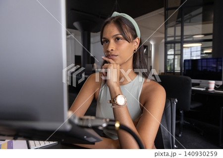 Adult asian woman sitting at desk in office, gazing at computer monitor with coffee cup 140930259