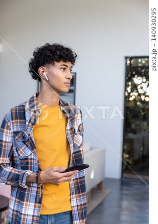 Man standing in modern living room holding smartphone and wearing earbuds looking right 140930295