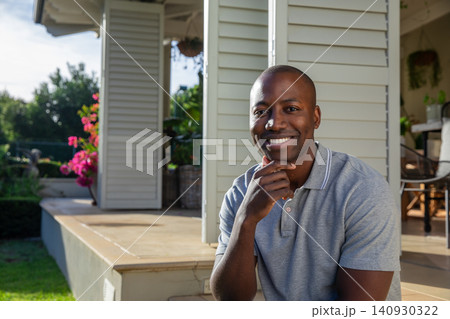 African American man sitting on patio step by white shutters in backyard with pink flowering plants African American man sitting on patio step by white shutters in backyard with pink flowering plants 140930322