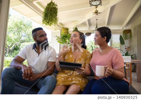 Diverse friends sitting on sofa under covered porch, sharing tablet and ceramic mug by garden Diverse friends sitting on sofa under covered porch, sharing tablet and ceramic mug by garden 140930325